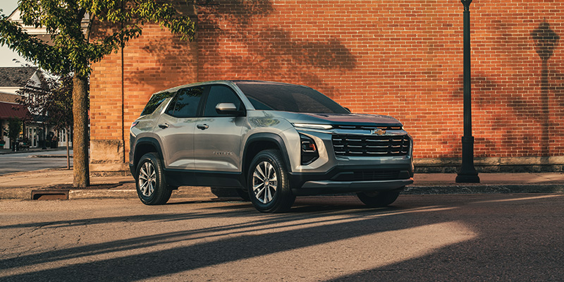 Silver Chevrolet Equinox SUV parked on city street near brick wall under sunlight